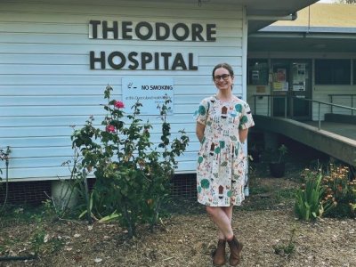 A woman in a dress and boots stands in front of a building which is labelled Theodore Hospital